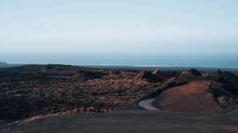 Wide landscape shot of an arid volcanic terrain in Lanzarote. Textured reddish-brown earth fills the foreground, transitioning into rugged black lava fields in the mid-ground. A grey winding road curves through the dark landscape. In the distance, the calm blue ocean meets a clear, gradient blue sky on the horizon.