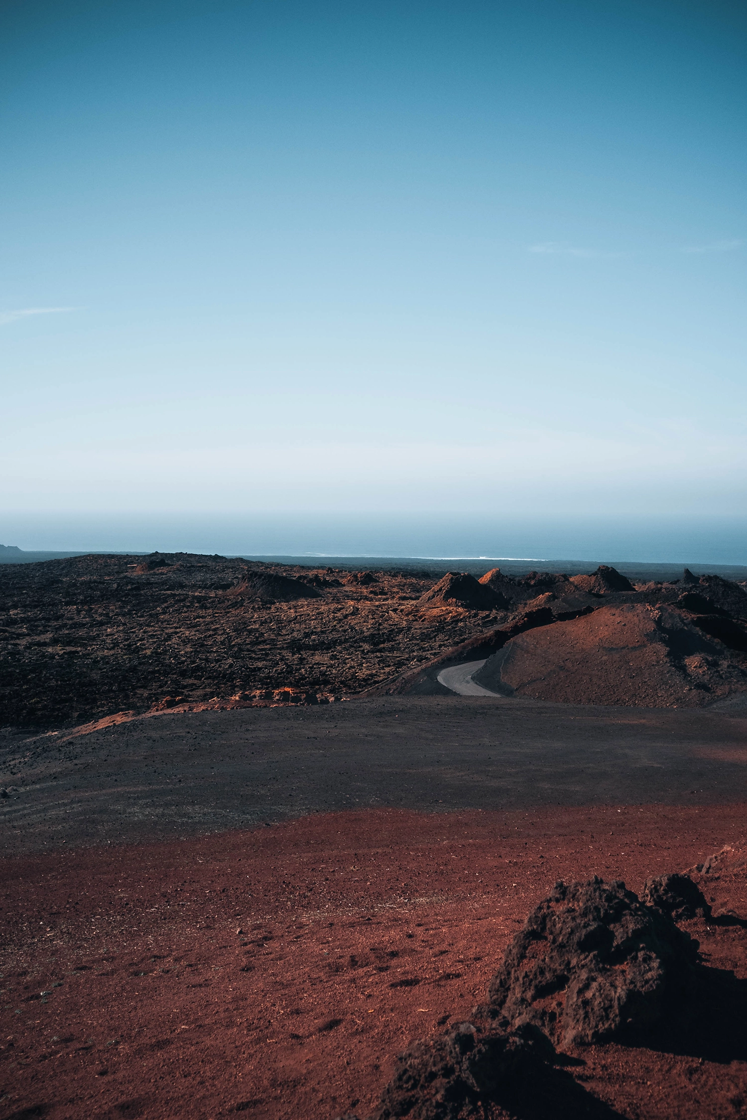 Wide landscape shot of an arid volcanic terrain in Lanzarote. Textured reddish-brown earth fills the foreground, transitioning into rugged black lava fields in the mid-ground. A grey winding road curves through the dark landscape. In the distance, the calm blue ocean meets a clear, gradient blue sky on the horizon.