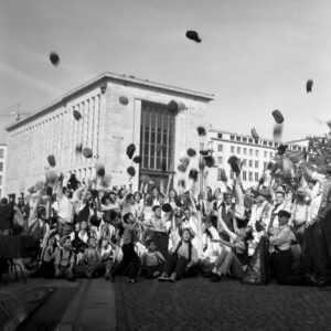 Black and white wide shot of a large crowd joyfully throwing hats and caps into the air in front of the Royal Library during the Zinneke Parade finale in Brussels, 2010.