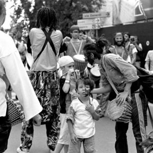 Black and white street photography of a percussion group with face paint playing drums, while a woman interacts with a young participant and a dog during the Par Tòt Parata 2007 in Bologna.