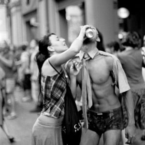 Black and white candid photo of a woman helping a shirtless participant wearing a tie drink from a cup during the festivities of the Par Tòt Parata 2007 in Bologna.