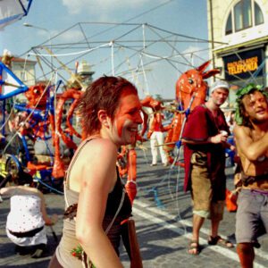 Color photograph of a smiling participant with red face paint walking past large red articulated puppets during the Par Tòt Parata 2007 festival in Bologna, Italy.