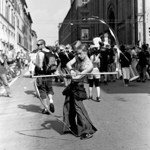 Black and white street photography of a bare-chested performer manipulating a long stick with ribbons attached during the Par Tòt Parata 2007 festival in Bologna.