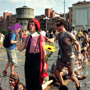 olor photograph of street performers, including one dressed as a mime holding a large sunflower and a small purple umbrella, interacting on a paved street during the Par Tòt Parata 2007 festival in Bologna, Italy.