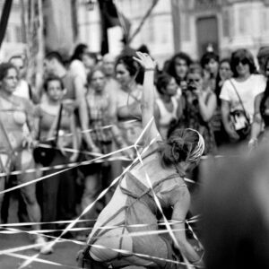 Black and white photograph of a street performer entangled in white ribbons during a dynamic performance in front of a crowd at the Par tòt Parata parade in Bologna, Italy, 2007.