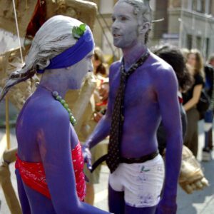 Two performers with purple-painted skin standing together at the Par tòt Parata in Bologna. The person on the left wears a red bandeau top and a blue headband, while the person on the right is shirtless, wearing a dark necktie, white shorts, and small prosthetic horns on their head. A large float structure made of straw is partially visible in the background.