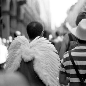 Rear view black and white photo of a participant wearing large white feathered wings walking through the crowd during the Par tòt Parata parade in Bologna, Italy, 2007.
