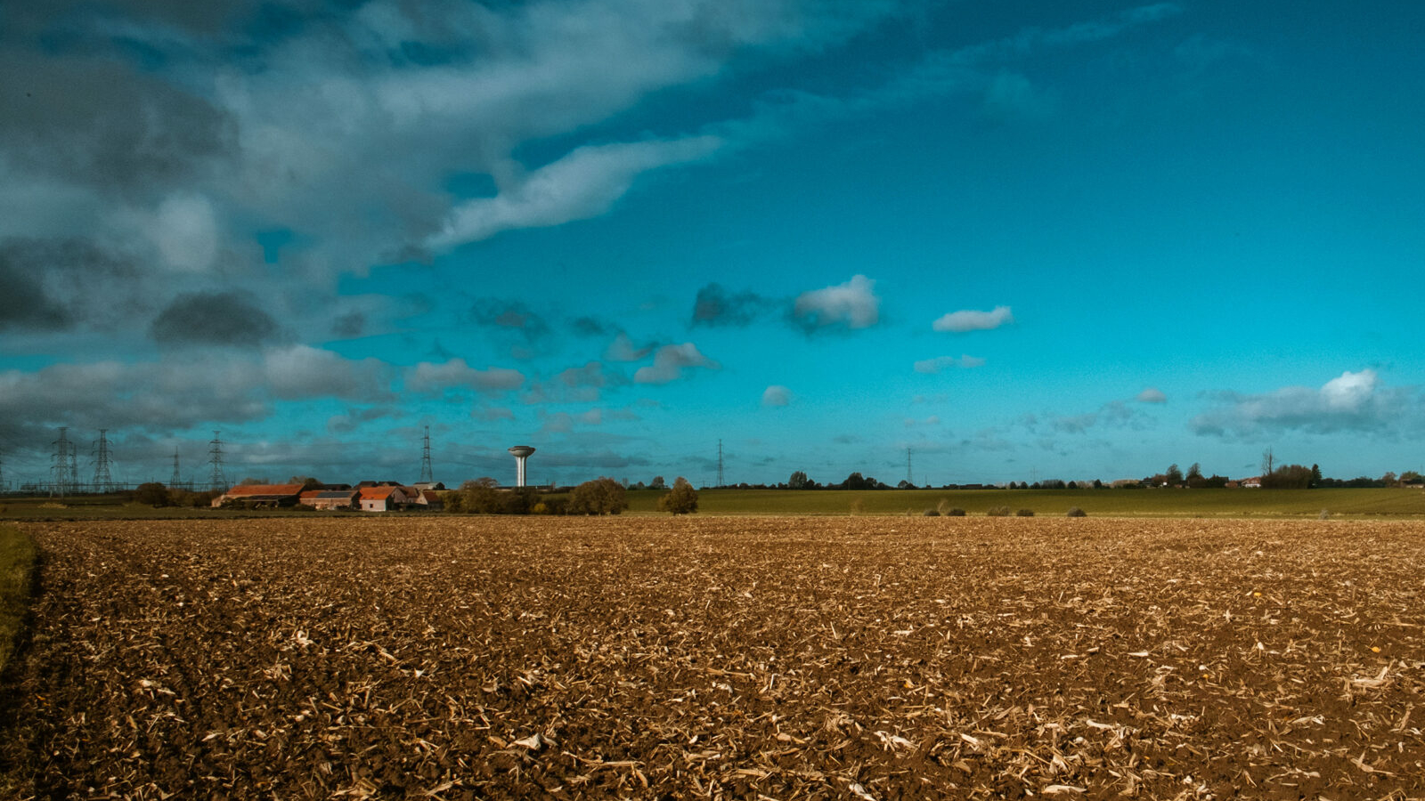 Expansive brown ploughed field stretching to the horizon under a vibrant blue sky with white clouds, featuring distant farm buildings and a water tower.