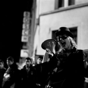 Black and white photograph of a woman performing at night during the Nuit Blanche festival in Brussels. She is wearing a top hat, sunglasses, and a spiked collar, shouting with her mouth wide open while holding a megaphone in one hand and a cigarette in the other. A blurred crowd and a building with a vertical Bosch sign are visible in the background.
