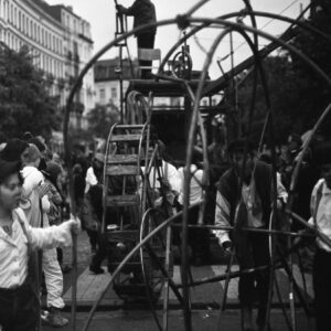 Black and white photo of participants maneuvering a large, complex metal wheel structure with a person perched on top during the Zinneke Parade 2010 in Brussels.