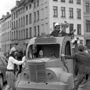 Black and white photo of a large DIY cardboard fire truck float labeled "POMPIERS" being pushed by participants on a cobblestone street during the Zinneke Parade 2012 in Brussels.