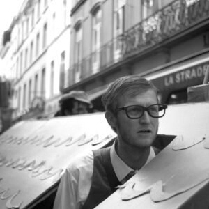 Black and white close-up photo of a participant wearing thick-rimmed glasses looking out from a cardboard house structure during the Zinneke Parade 2012 in Brussels streets.