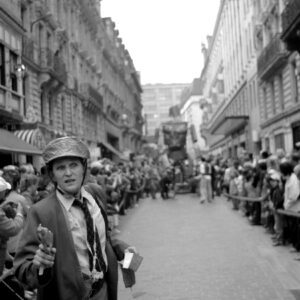 Black and white photo of a performer wearing a suit and a makeshift helmet holding a prop, walking down a crowded street with floats in the background during the Zinneke Parade 2012 in Brussels.
